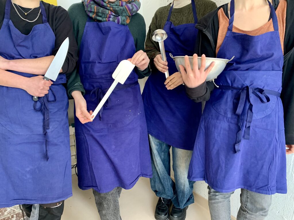 Phot en couleur de 4 personnes bénévoles et salariées membres de l'association Coco Cantine qui portent des tabliers bleus de cuisine et tiennent à la main des ustensiles pour fabriquer des plats de saison et bons : couteau, maryse, louche et saladier. On ne voit pas leurs visages.
