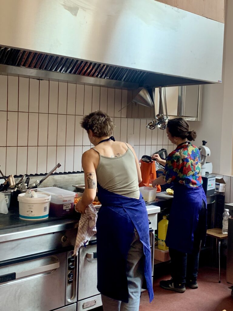 Photo intérieur jour en couleur, qui illustre l'investissement bénévole associatif dans la cuisine du restaurant végétarien Coco Cantine. On y voit 2 personnes, de dos, qui préparent un plat végan dans une cuisine professionnelle. Elles portent des tabliers bleus.