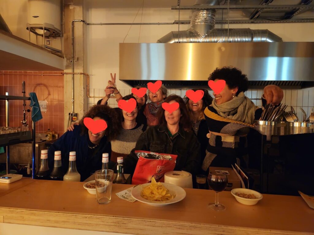 Photo couleur prise en intérieur de nuit. 7 personnes dont le visage est anonymisé par des coeurs rouges posent en souriant derrière le comptoir. Cette image illustre la mission de bénévolat pour le bar de la Coco Cantine, lieu convivial engagé pour l'accessibilité du bien manger et l'alimentation durable dans le 20ème arrondissement de Paris.