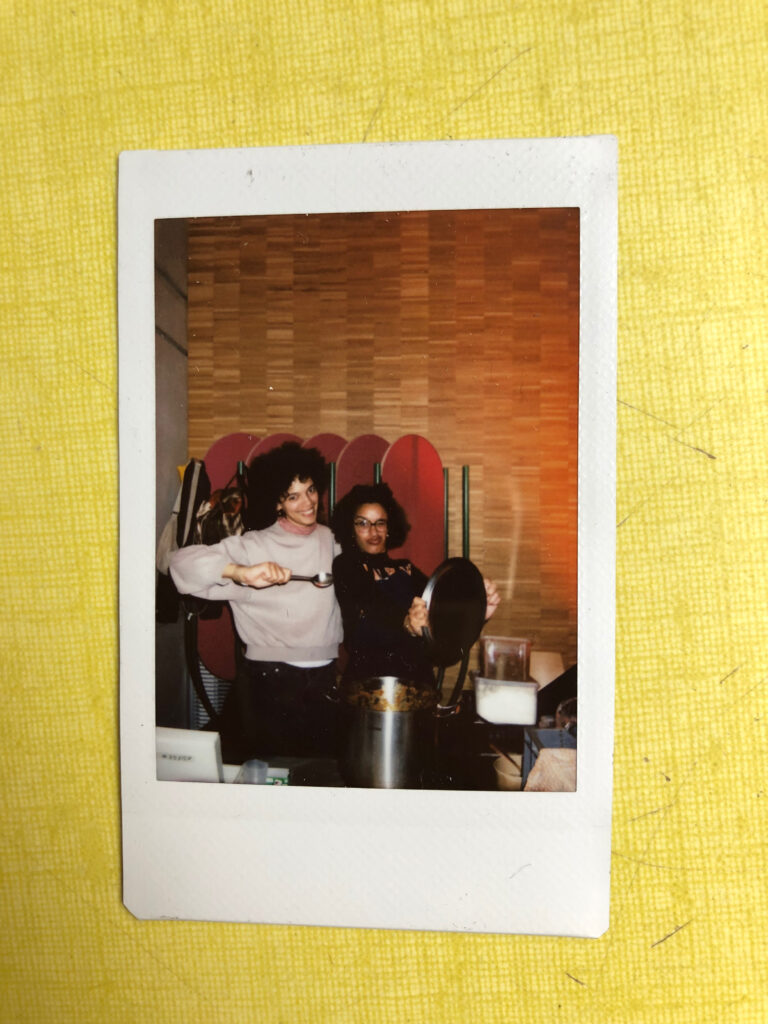 Polaroid couleur des membres de la cantine associative au catering d'une prestation traiteur pour une dramaturge/metteuse en scène lors d'un événement culturel dans le 3ème arrondissement parisien. On y voit 2 personnes à la peau marron, cheveux frisés, de taille différentes, posant avec une grande marmite de rougail, tenant dans leurs mains le couvercle de la marmite et une cuillière de service. La photo est posée sur une surface en formica jaune.